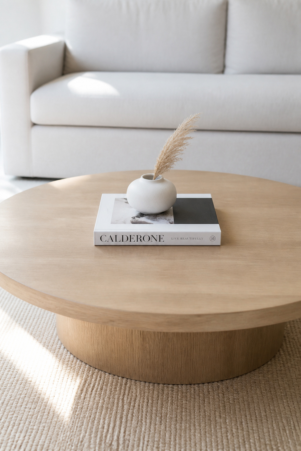 A pale round oak coffee table with a single large white art book placed flat in the centre and a small white ceramic vessel on top, surrounded by clear negative space, illustrating the single-book minimalist coffee table book styling approach