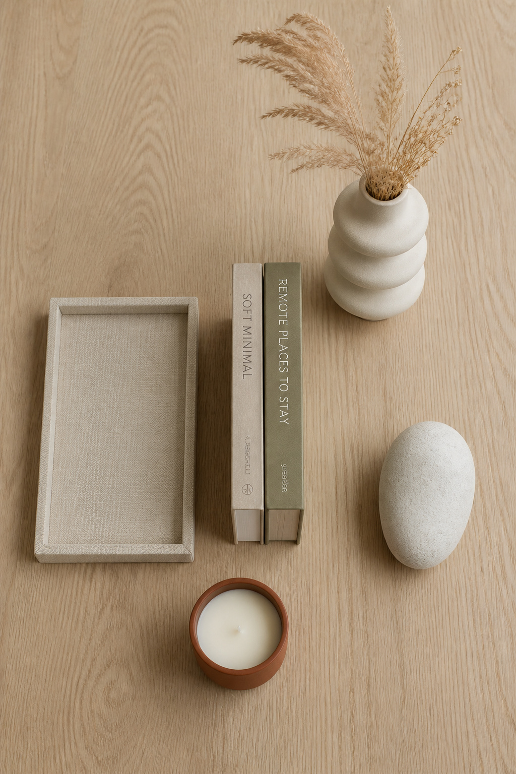 Five coffee table styling objects arranged on oak surface: linen tray, stacked books, tall ceramic vase with dried grasses, terracotta candle and smooth stone accent