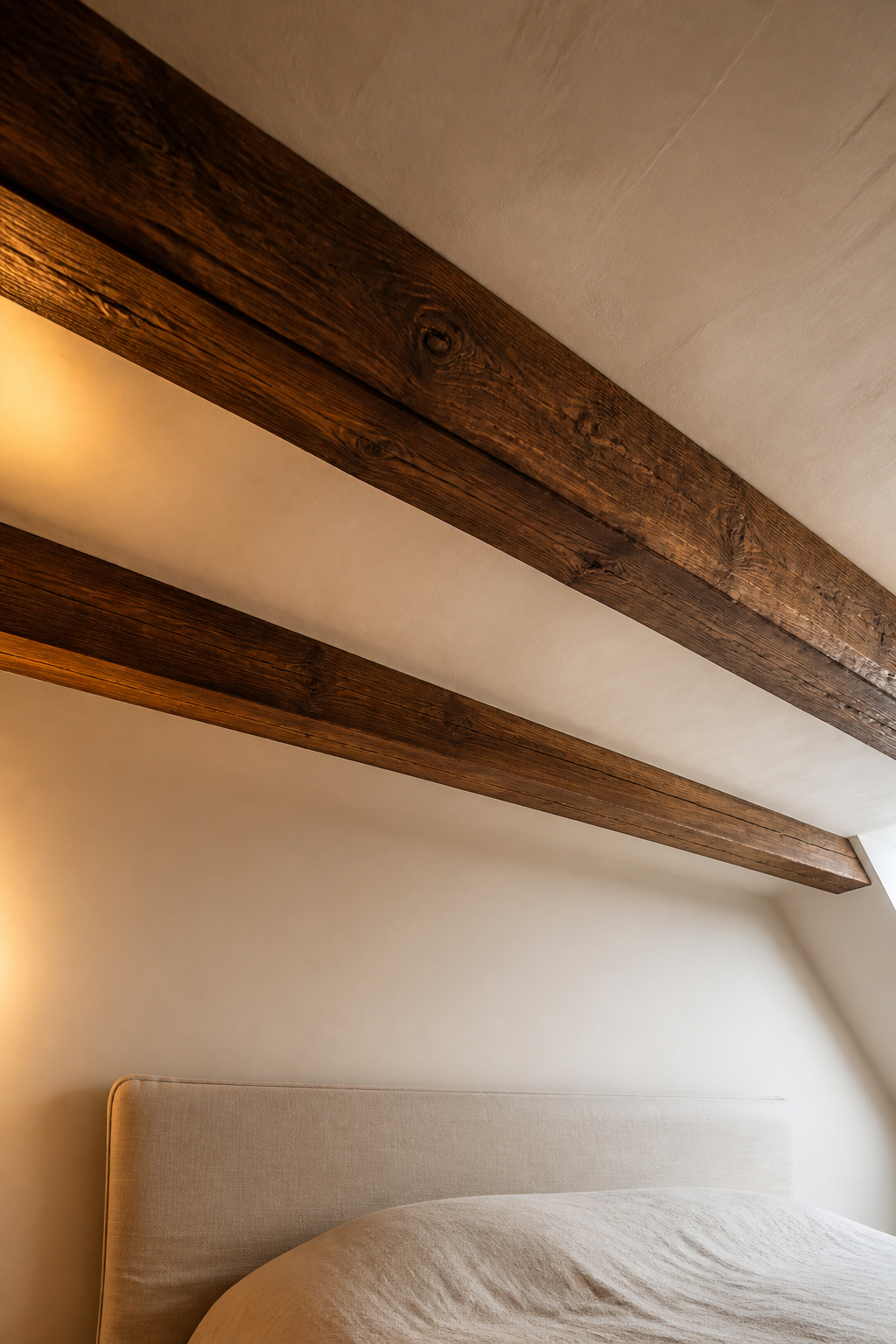 Two dark-stained exposed timber beams crossing a warm white plastered attic ceiling, grain visible, casting warm shadows with amber spotlight accent