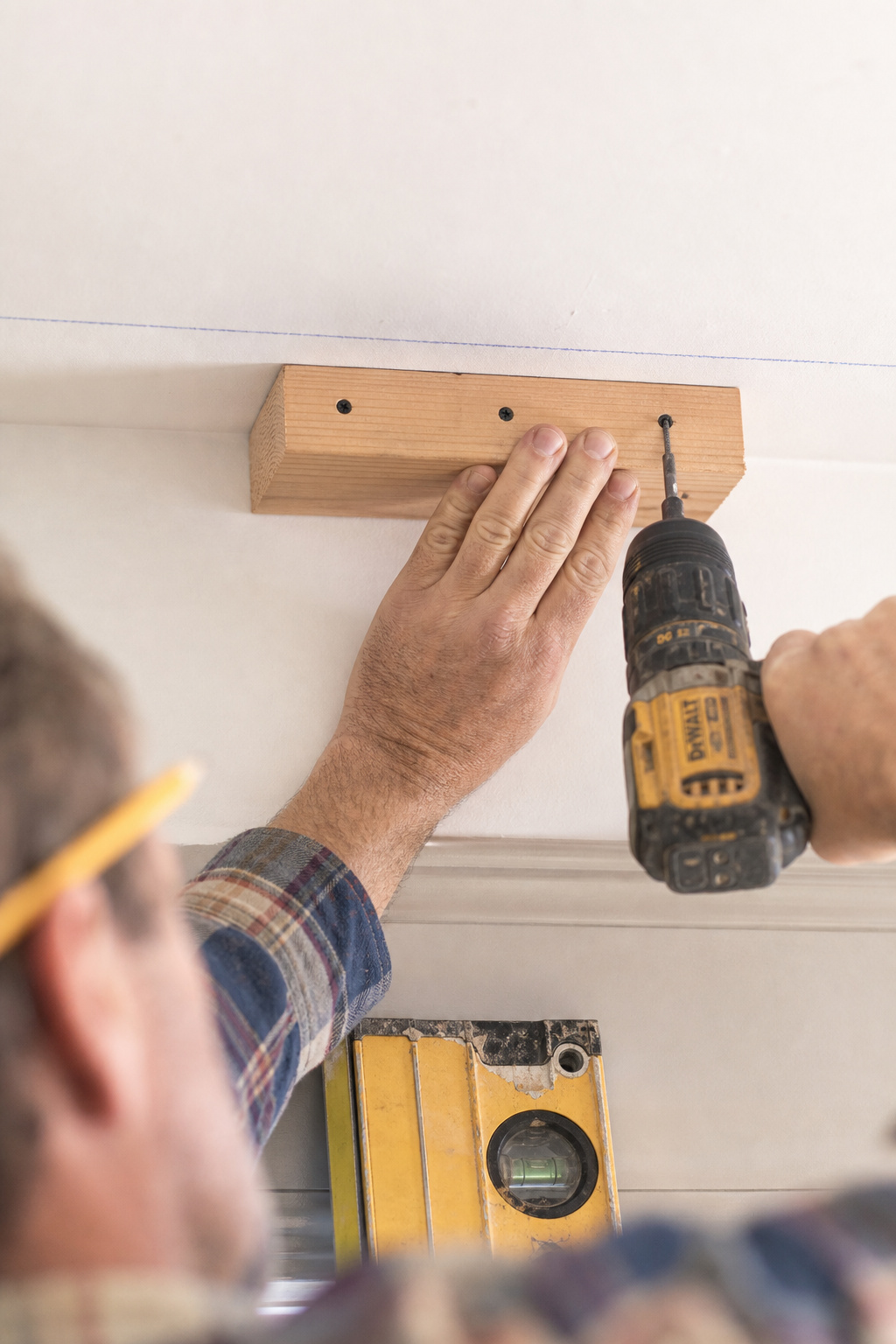 Installer screwing a 2x4 nailer block to the ceiling along a chalk line, drill bit visible, as the first step in faux beam installation