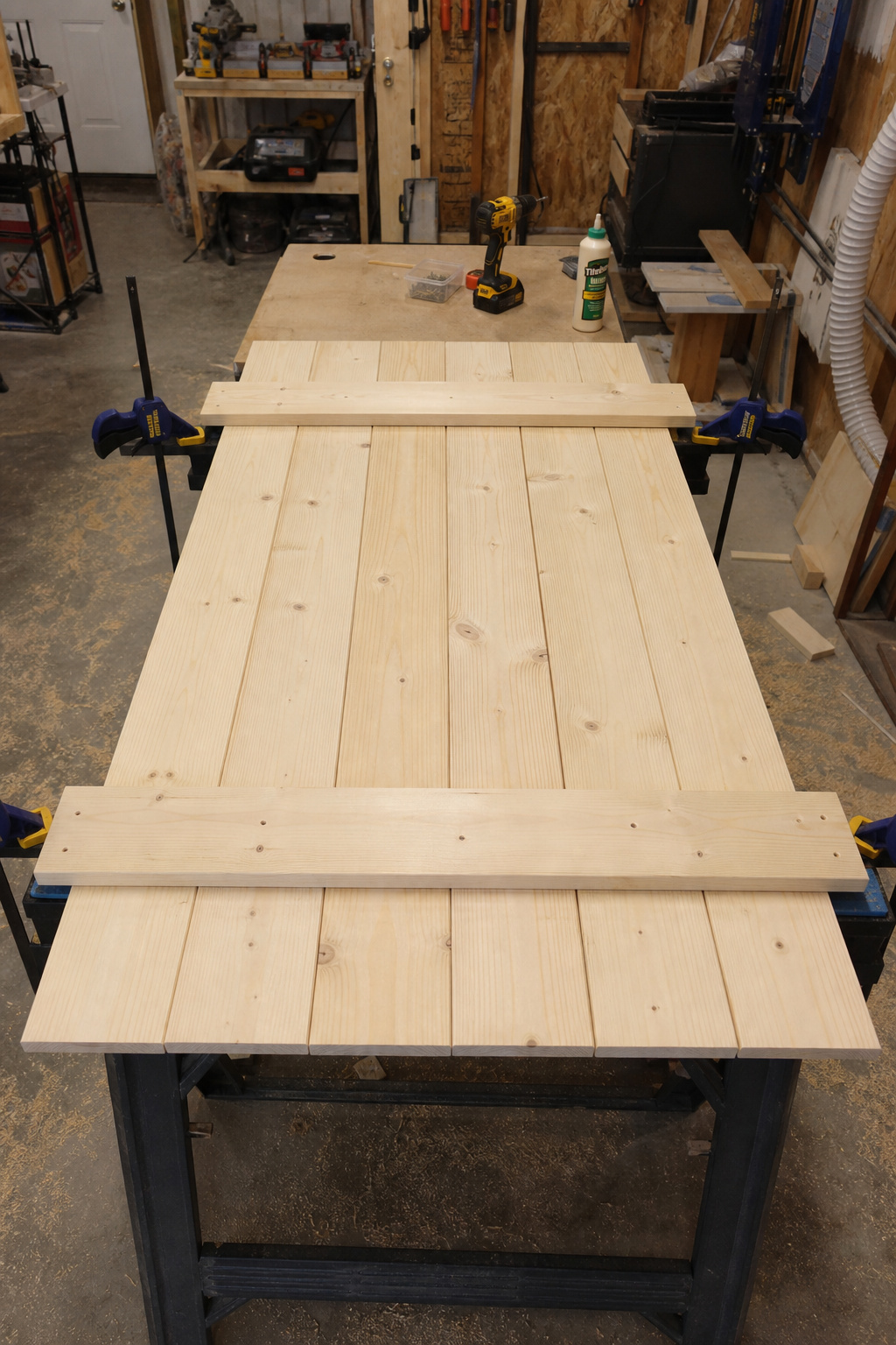 DIY barn door being assembled on sawhorses — pine planks with horizontal battens, drill and clamps in a home workshop