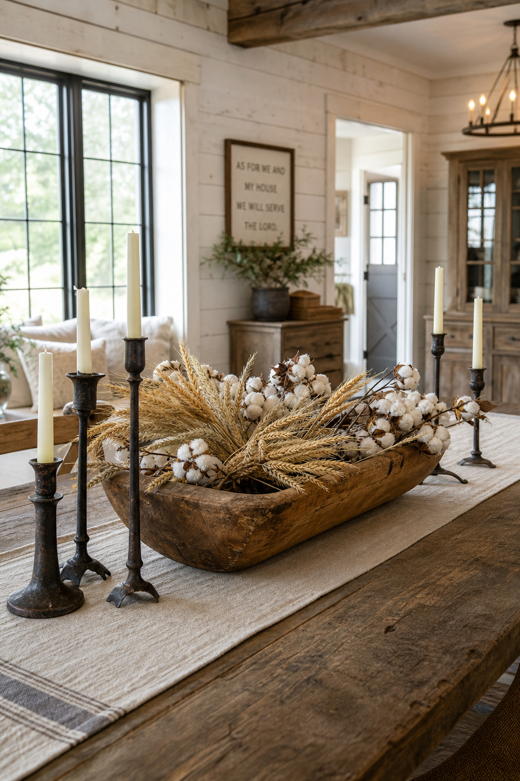 Long reclaimed-wood farmhouse dining table styled with a wooden dough bowl centerpiece, iron taper candlesticks, dried wheat stems, and a natural linen runner