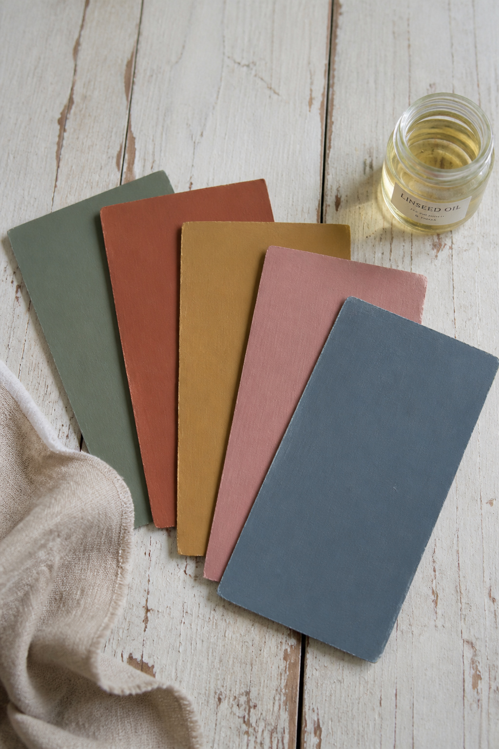 Five heritage paint chips in muted sage, rust, ochre, dusty rose and slate blue arranged on aged timber beside a jar of linseed oil