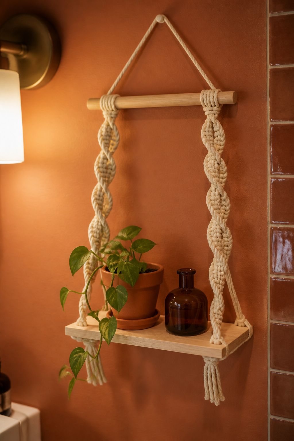 Finished DIY macrame hanging shelf on a terracotta bathroom wall holding a trailing pothos and amber glass bottle