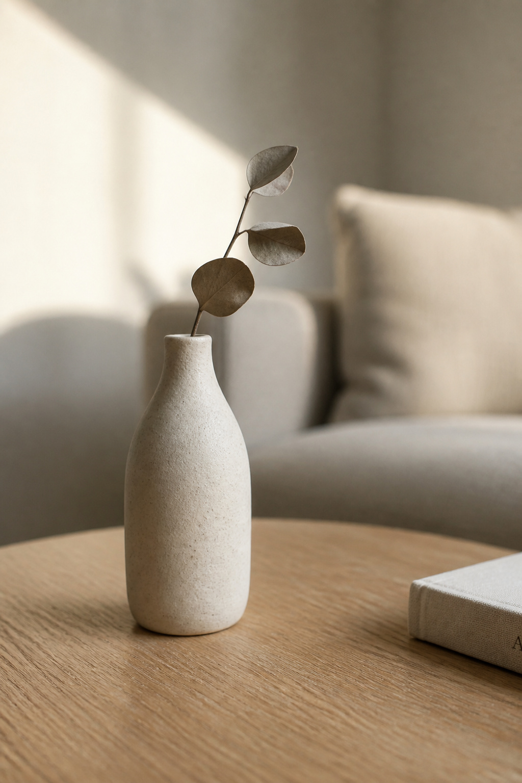 A slim unglazed stoneware bud vase with a single dried eucalyptus stem on a light oak coffee table, representing the living element in a minimalist coffee table arrangement