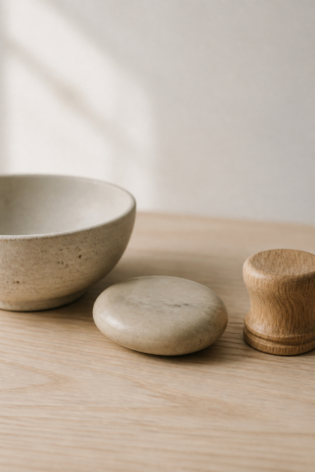 Close-up of three natural material objects on an ash wood surface — a matte unglazed stoneware bowl, a honed limestone oval, and a small turned oak object — illustrating the texture-based visual interest of natural minimalist materials