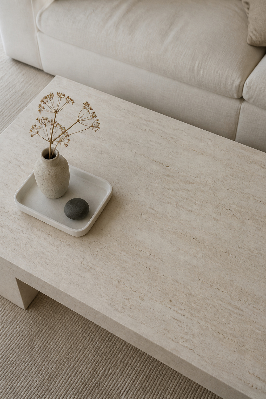A pale stone rectangular coffee table with a small minimalist vignette in the left third — a white ceramic tray, stoneware vase, and dark river stone — and the right two-thirds completely bare, demonstrating the negative space principle in minimalist coffee table decor