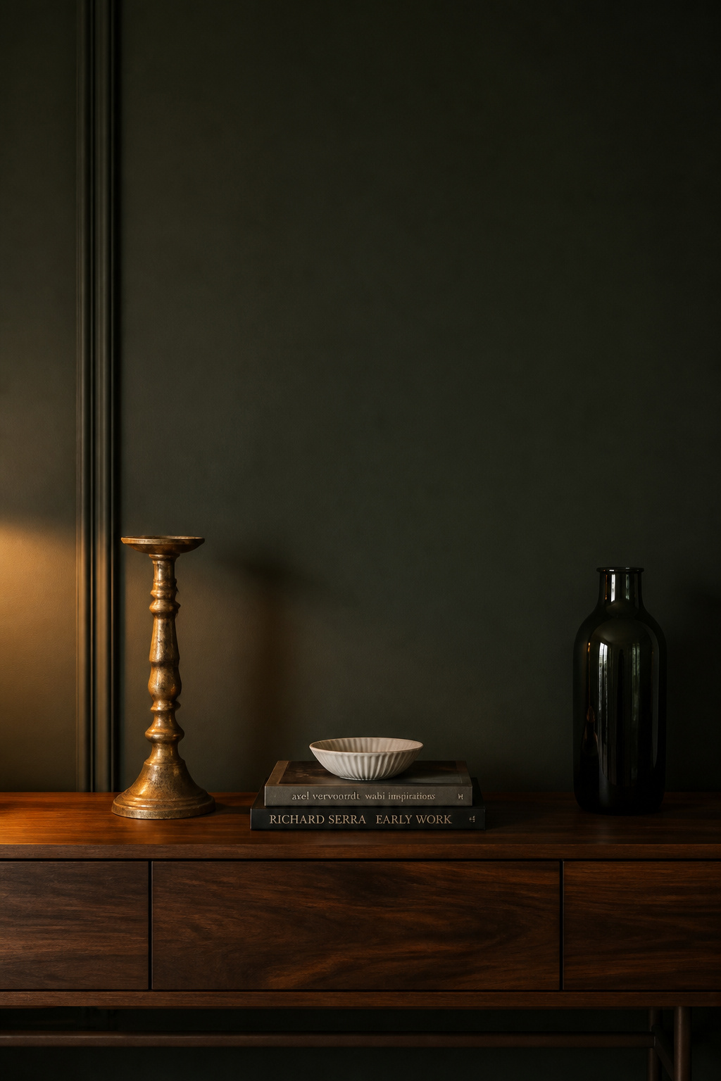 Three curated objects — aged brass candlestick, art book stack with white ceramic, and dark glass vase — against deep green wall showing the collected styling standard for moody living rooms