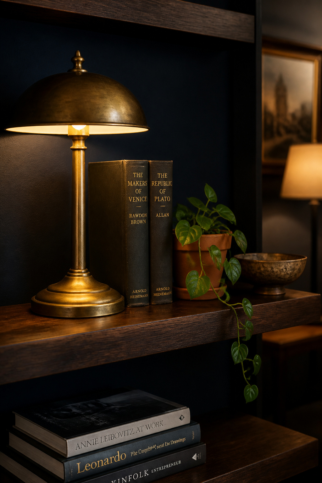 Dark walnut shelf styled with brass lamp, stacked books, trailing plant and bronze bowl against deep navy wall