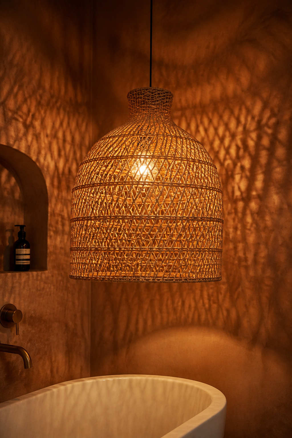 Rattan bell pendant lampshade hanging over a white freestanding bath, casting a latticed golden glow from warm 2700K light onto a terracotta limewashed wall