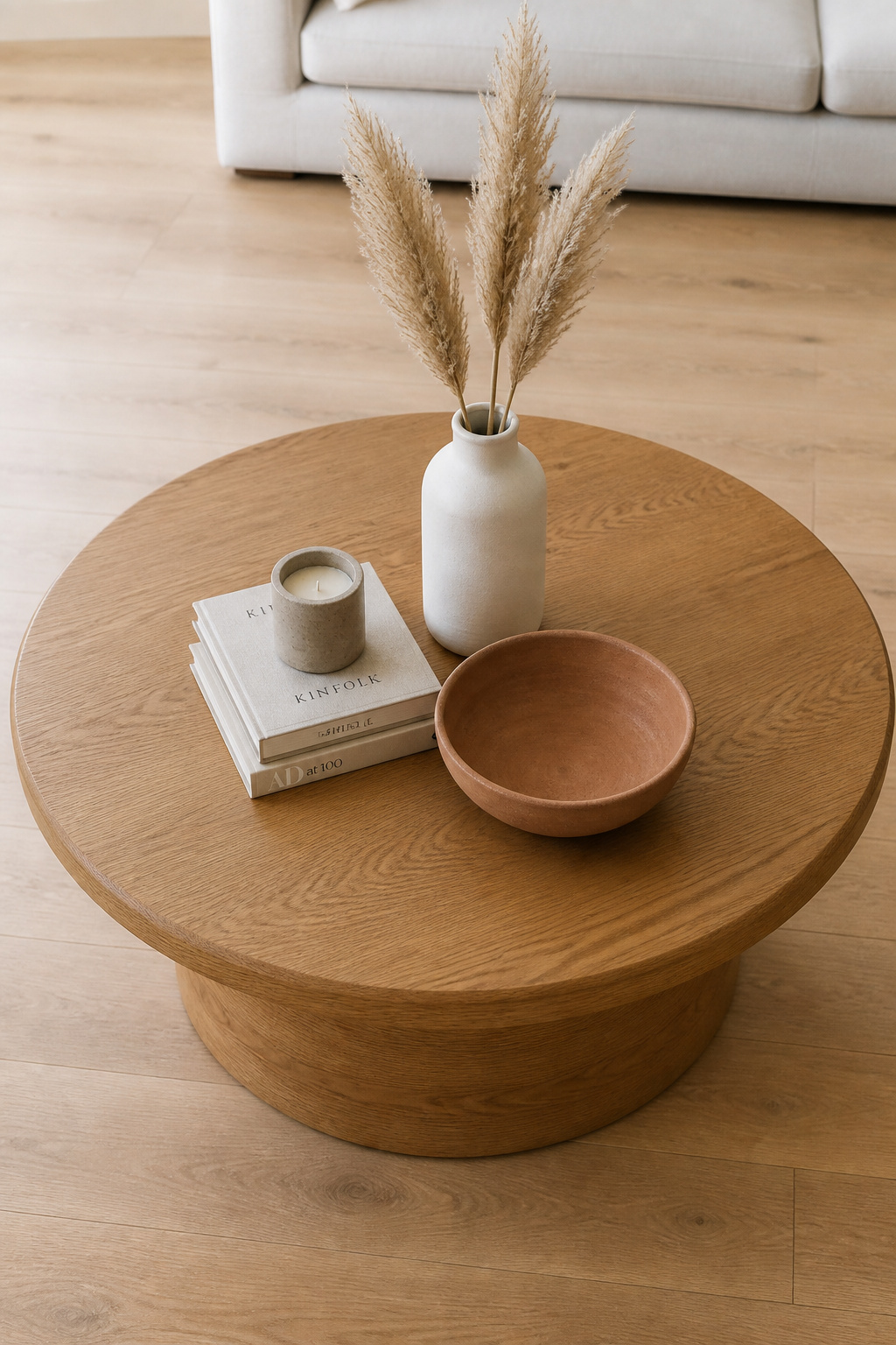 Round oak coffee table styled with a central triangle of matte ceramic vase with dried stems, stacked books, and a stoneware bowl