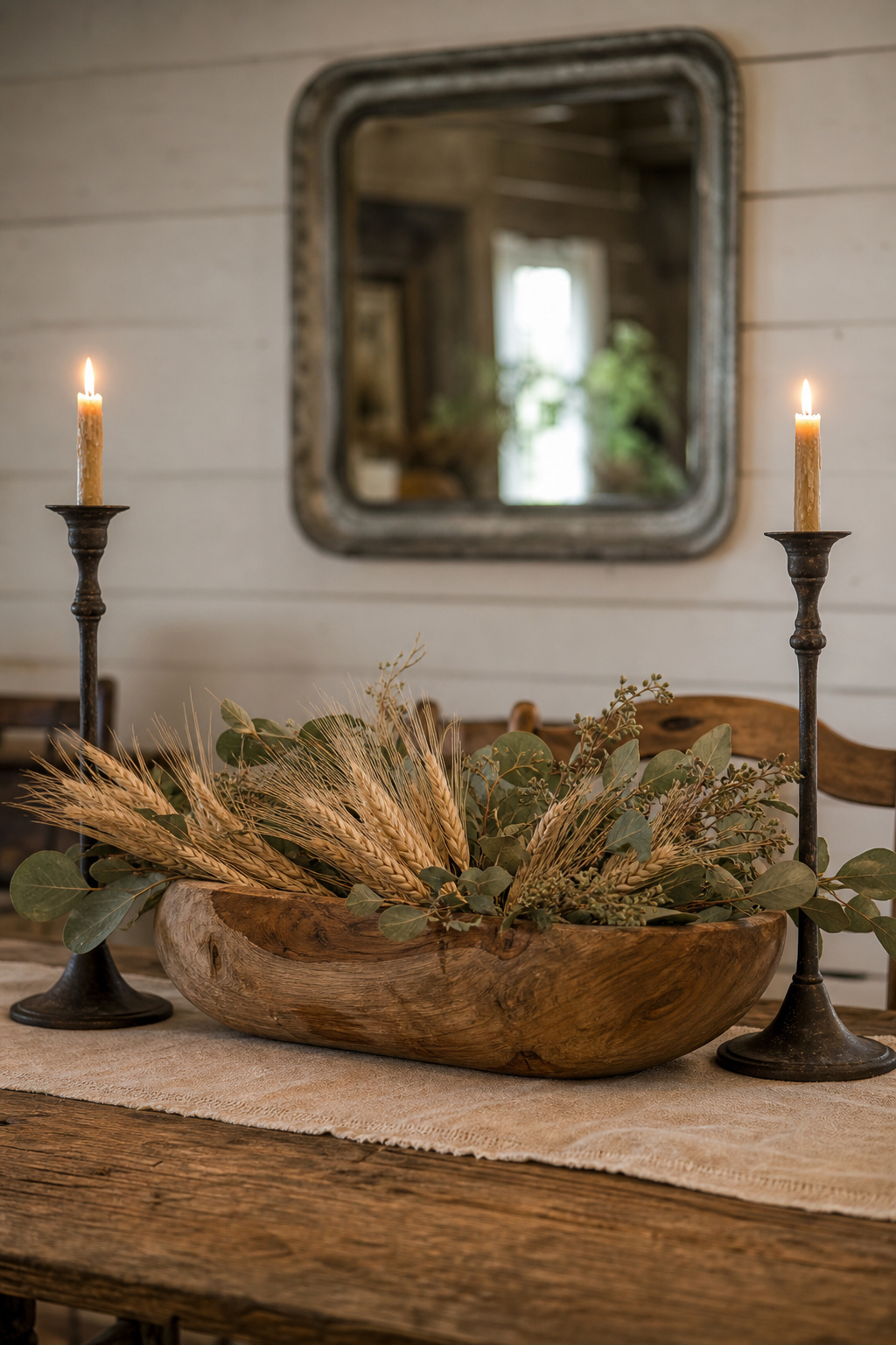 Rustic farmhouse dining table centrepiece with wooden bowl holding dried wheat and eucalyptus, flanked by iron taper candlesticks on an unbleached linen runner