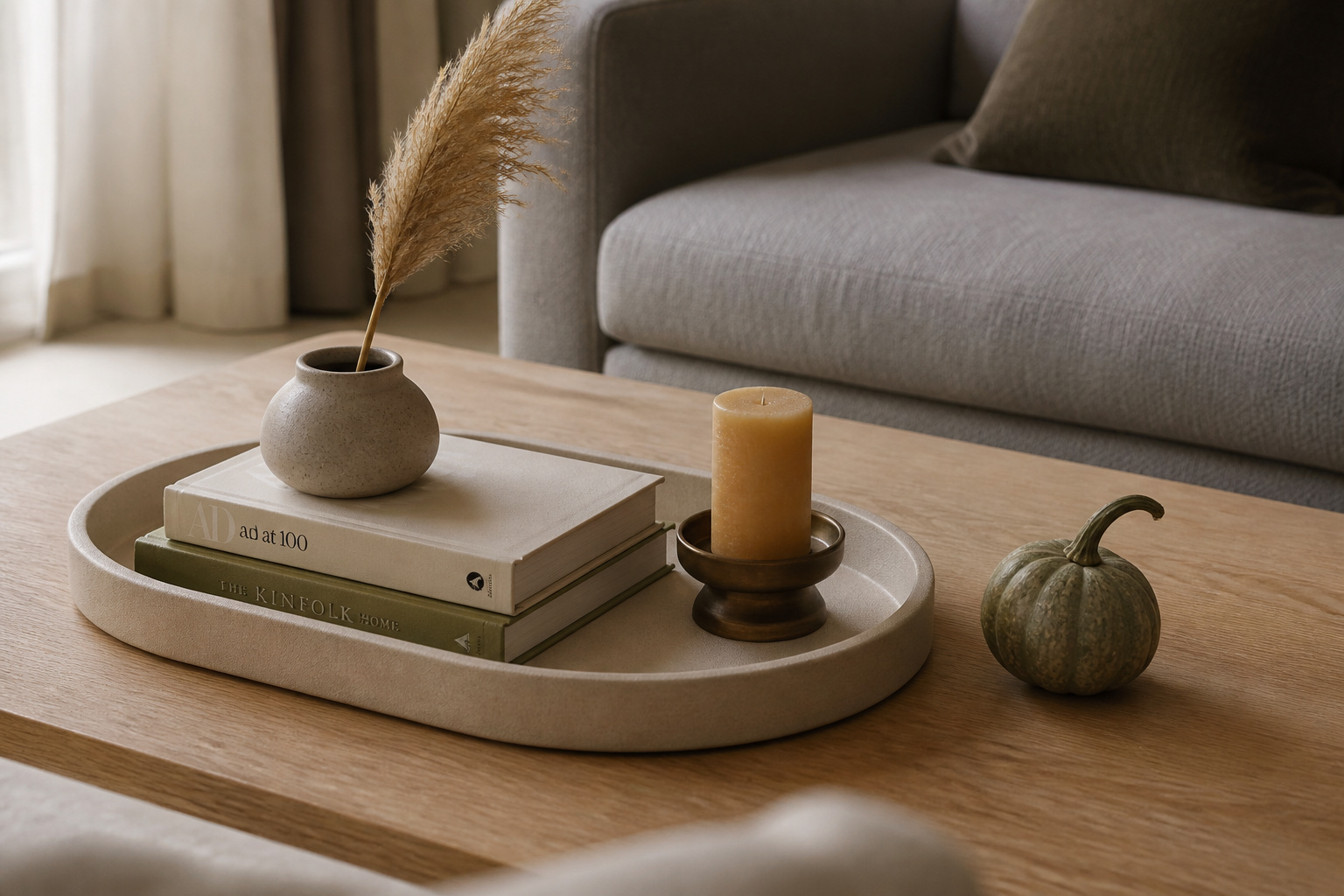 A neutral linen tray on a light oak coffee table styled with a stoneware vase, dried pampas stem, book stack, and bronze candle holder — the swappable-base approach to seasonal coffee table decor