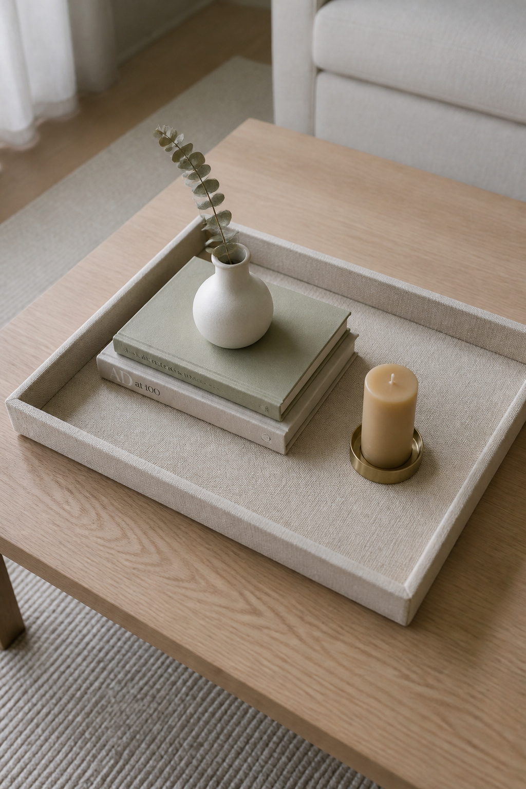 Overhead view of a neutral linen tray on a pale ash coffee table with a book stack, ceramic bud vase, and beeswax pillar candle — the year-round base for seasonal coffee table decor swaps