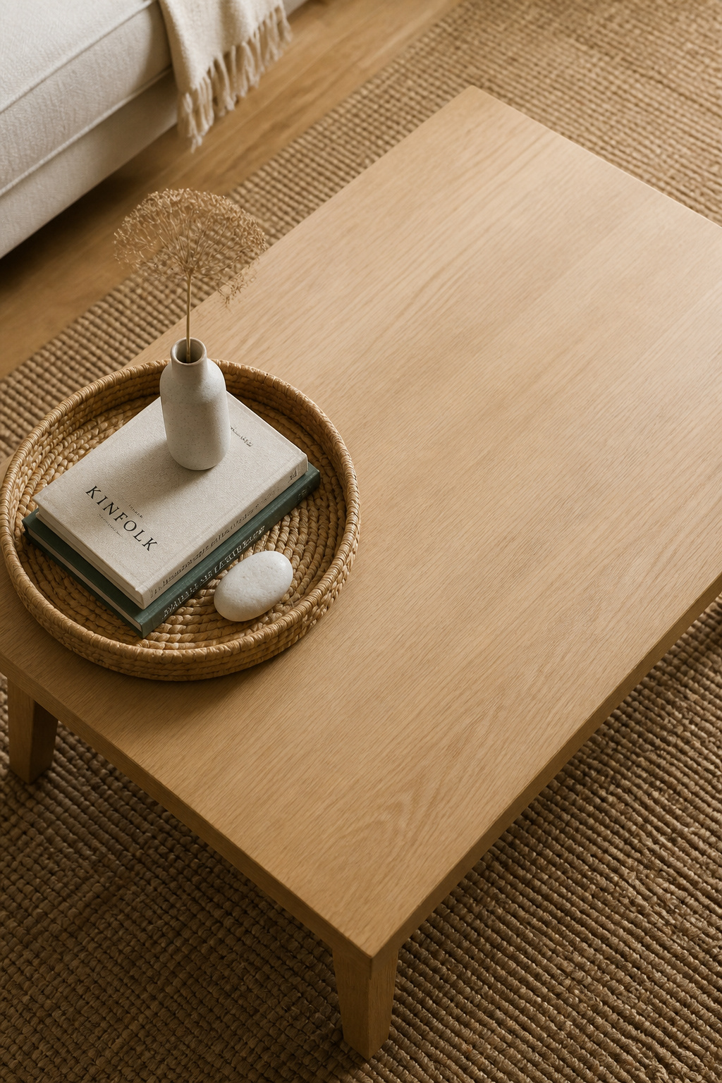 A compact rectangular coffee table with a single 12-inch woven tray vignette occupying the left third — two stacked books, a bud vase, and a smooth stone inside the tray — and the remaining two-thirds of the surface completely clear