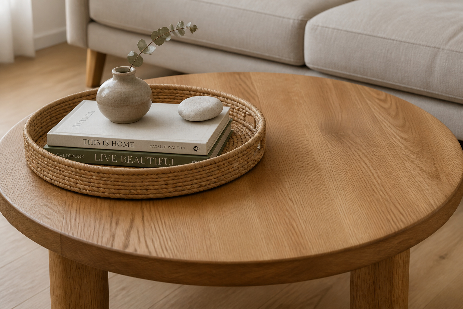 A compact round oak coffee table with one woven tray holding a small book stack, bud vase with eucalyptus, and a river stone — the opposite end of the surface completely clear — demonstrating small living room coffee table decor with one tight vignette