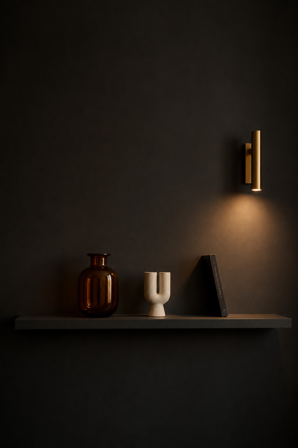 A wall-mounted shelf in matte charcoal matching the dark living room walls — three objects only with deliberate breathing room between them, lit by a single brass wall sconce