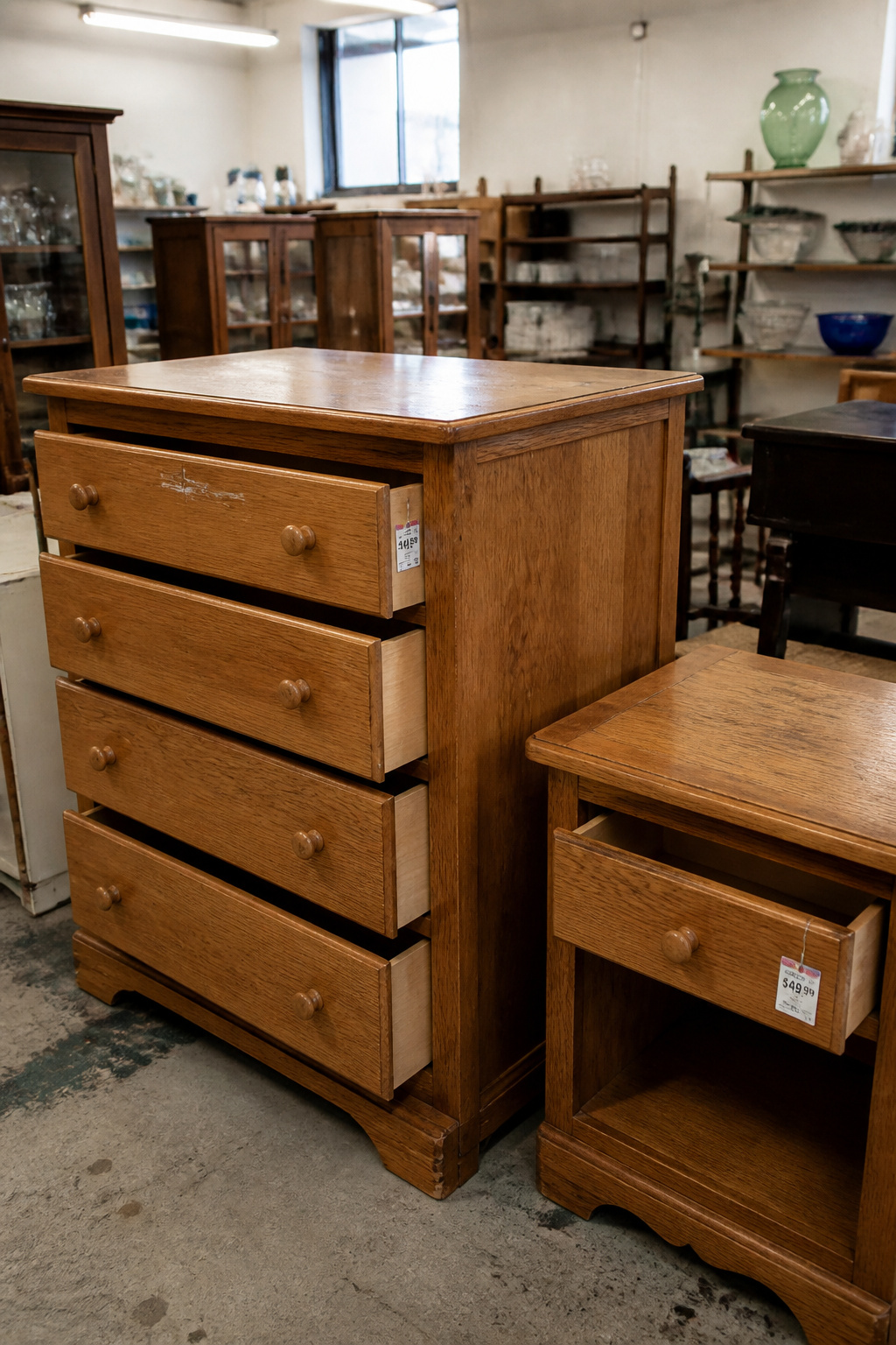A solid oak dresser and matching nightstand at a thrift store, drawers open showing clean interiors, both with visible honest wear and thrift store price tags