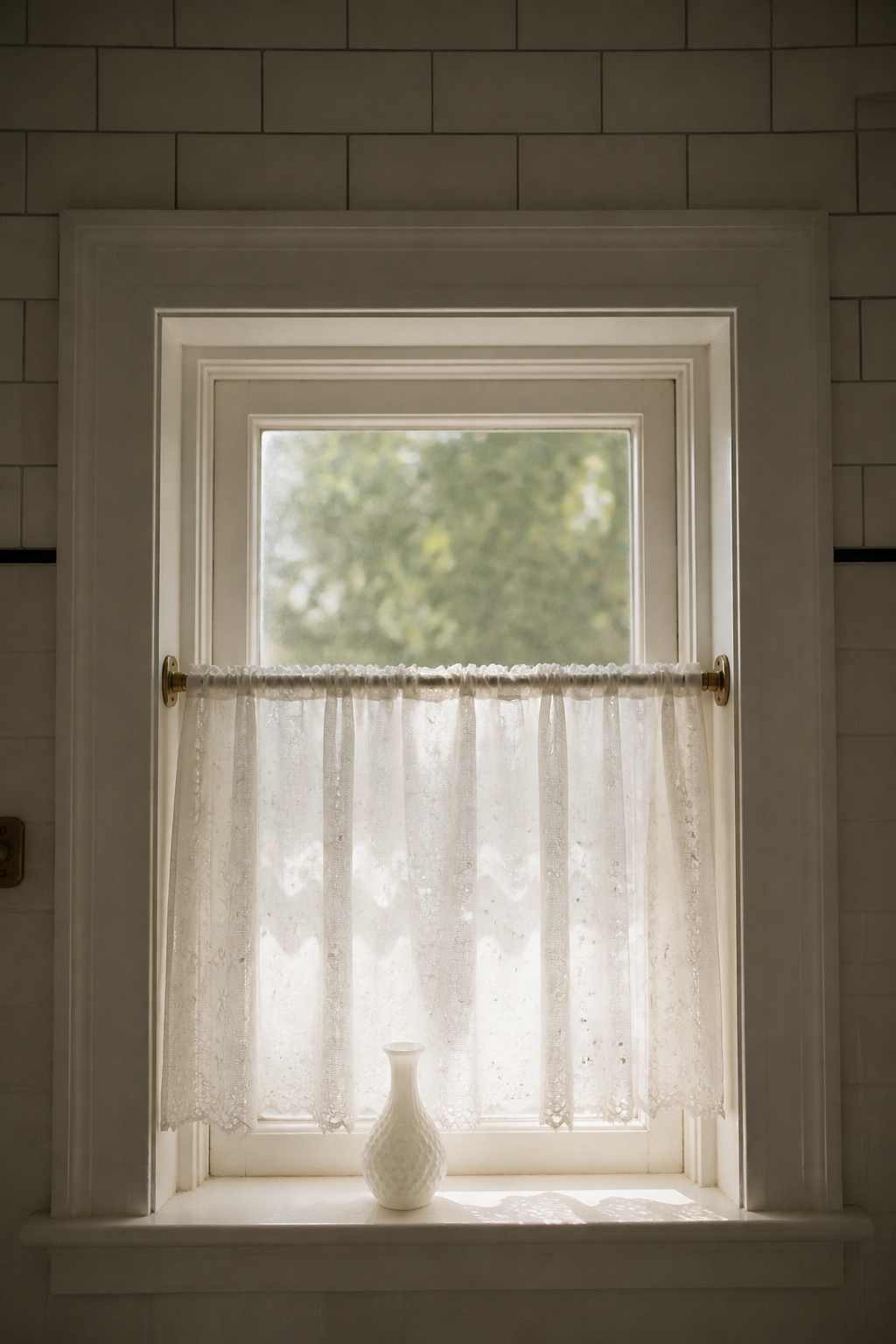 White cotton eyelet café curtains on a brass tension rod covering the lower half of a bathroom window, with soft morning light filtering through