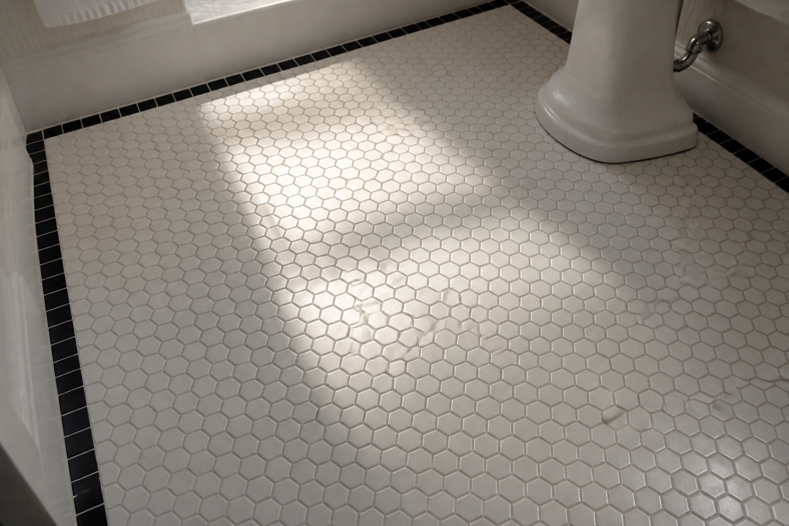 Overhead view of a period-accurate vintage bathroom floor with white 1-inch hex mosaic tiles and a black square border, grey grout lines