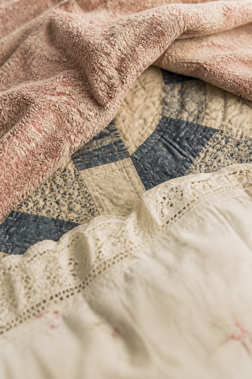 Close-up overhead view of layered vintage bed textiles — patchwork quilt, chenille, and lace pillowcase in warm morning light