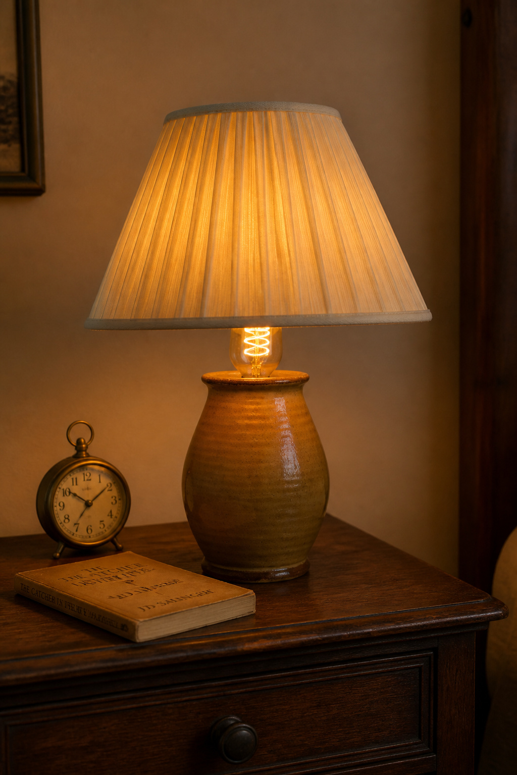 Vintage bedside lamp with hand-thrown ochre ceramic base and pleated ivory linen shade, Edison filament LED glowing amber inside, on a dark walnut nightstand
