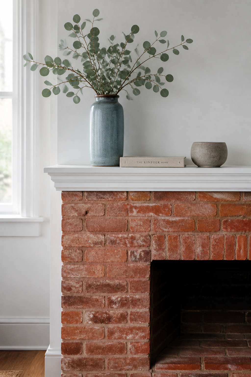 Red brick fireplace with white wooden mantel shelf holding dusty-blue ceramic vase, eucalyptus, and grey stoneware — cool spring tones contrasting deliberately against the warm brick