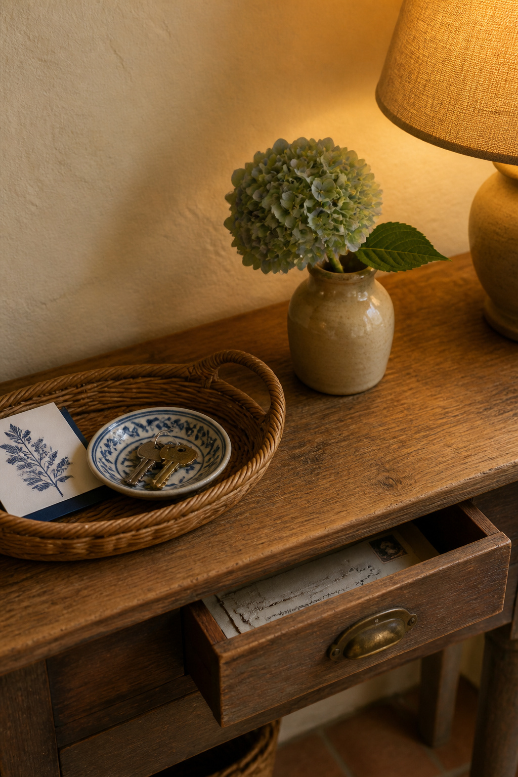 A cottage console table surface with a wicker tray holding a ceramic key dish, a single drawer holding mail, and a clear display surface with a lamp and hydrangea stem