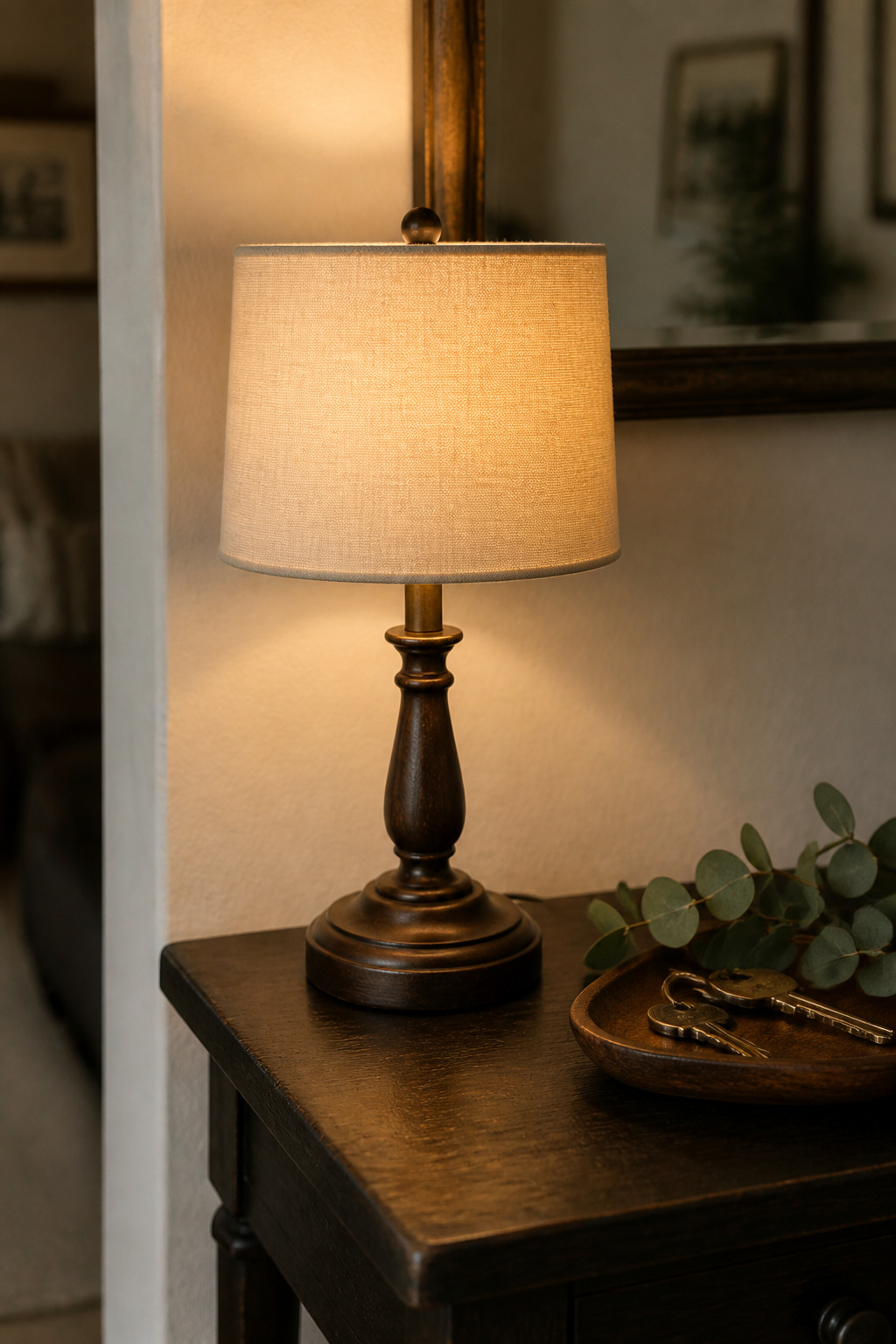A small cream linen-shaded table lamp on a dark narrow console table in a cottage entryway, casting warm amber light across the surface and surrounding a key tray and plant sprig