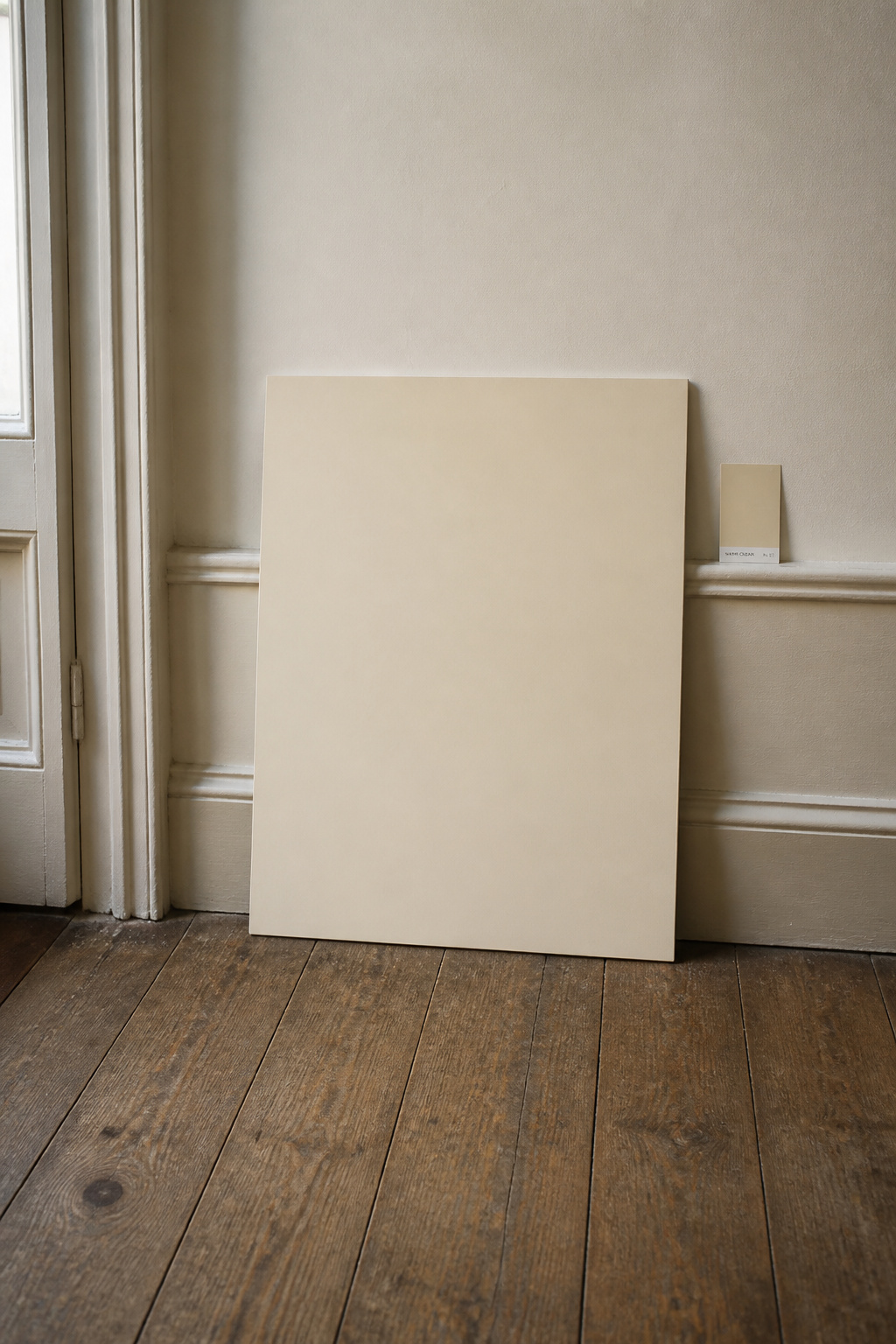 A large warm-cream paint sample board leaning against a period cottage entry wall at floor level, beside the skirting and on aged oak flooring — illustrating the professional large-sample testing method