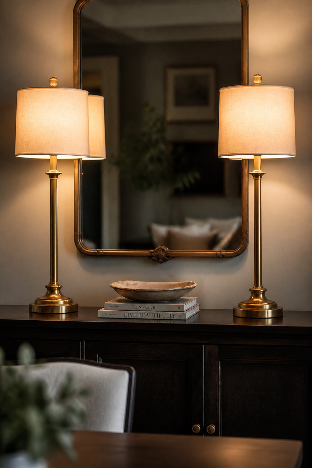A pair of slim brass buffet lamps with cream shades at the ends of a dark sideboard, flanking a ceramic bowl and stacked books at the centre