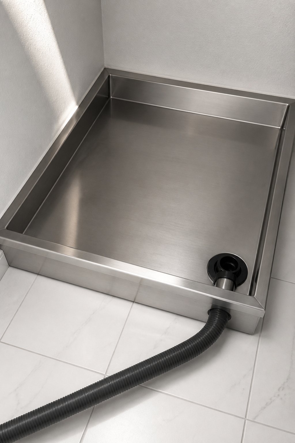 Stainless steel washing machine drain pan on white tile floor with 1-inch PVC drain fitting and drain hose routing to baseboard, pan seated tight against back wall