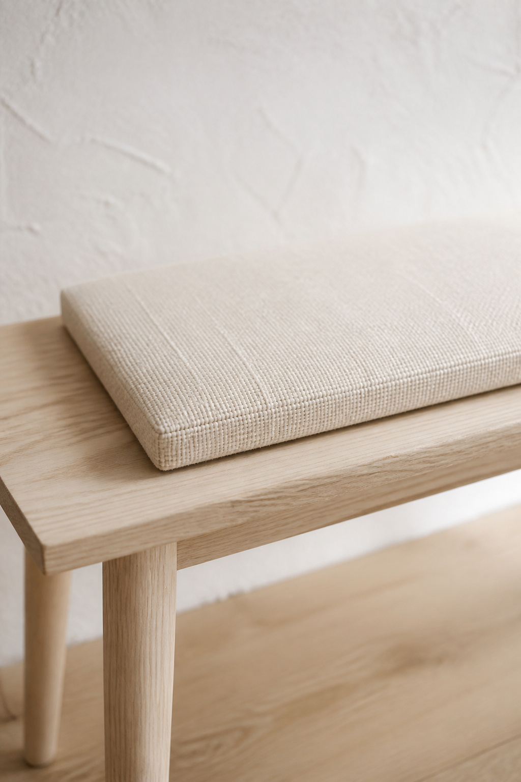 Close-up of a woven cream-linen bench cushion sitting at two-thirds seat width on a pale ash entryway bench, with bare wood visible at each end