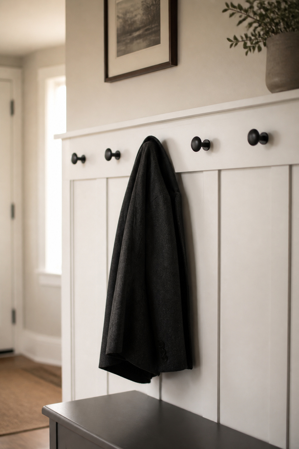 White shaker hook rail with five matte-black hooks at 60-inch height above an entryway bench, single wool coat hanging