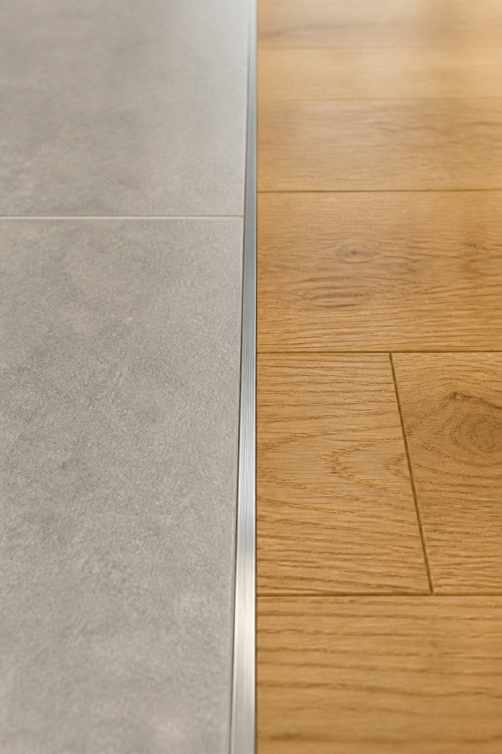 Close-up of a clean tile-to-hardwood floor transition using a slim brushed stainless Schluter profile, with warm grey porcelain tile on the left and honey-oak hardwood on the right