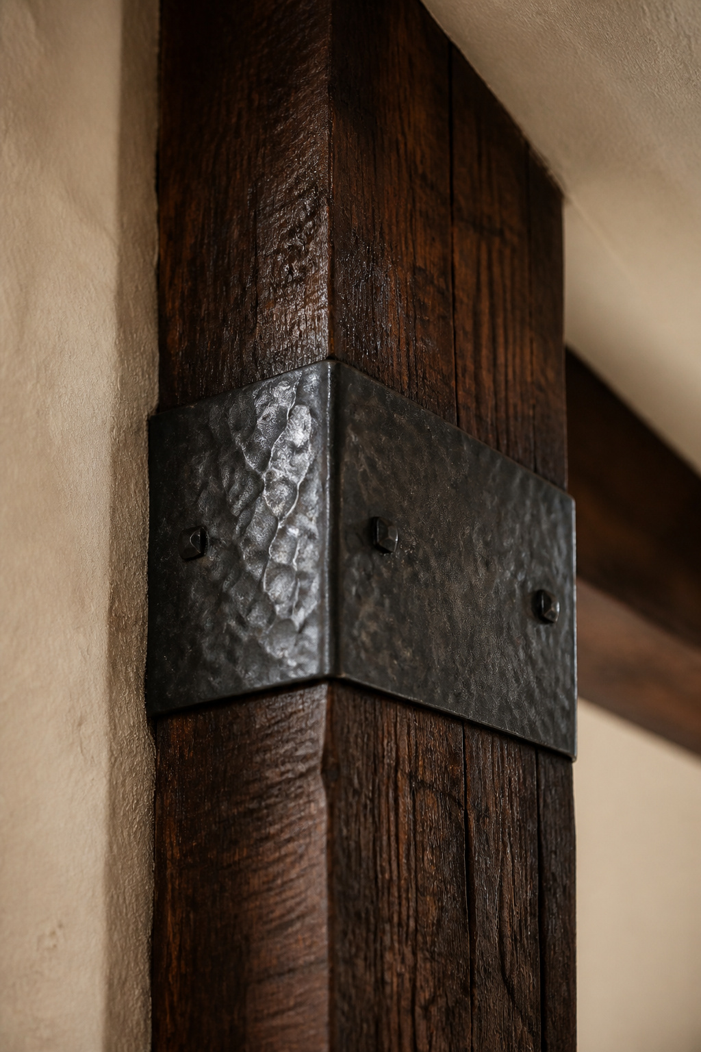 Dark walnut oak ceiling beam at plaster wall junction with a hand-forged wrought-iron strap wrapped around the beam, raking warm light on the iron texture