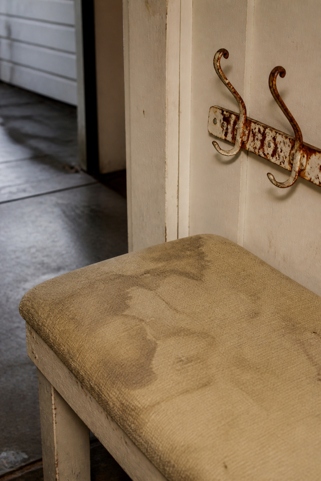 Water-damaged fabric bench and rusted painted hook at a garage entryway showing why interior furniture fails there