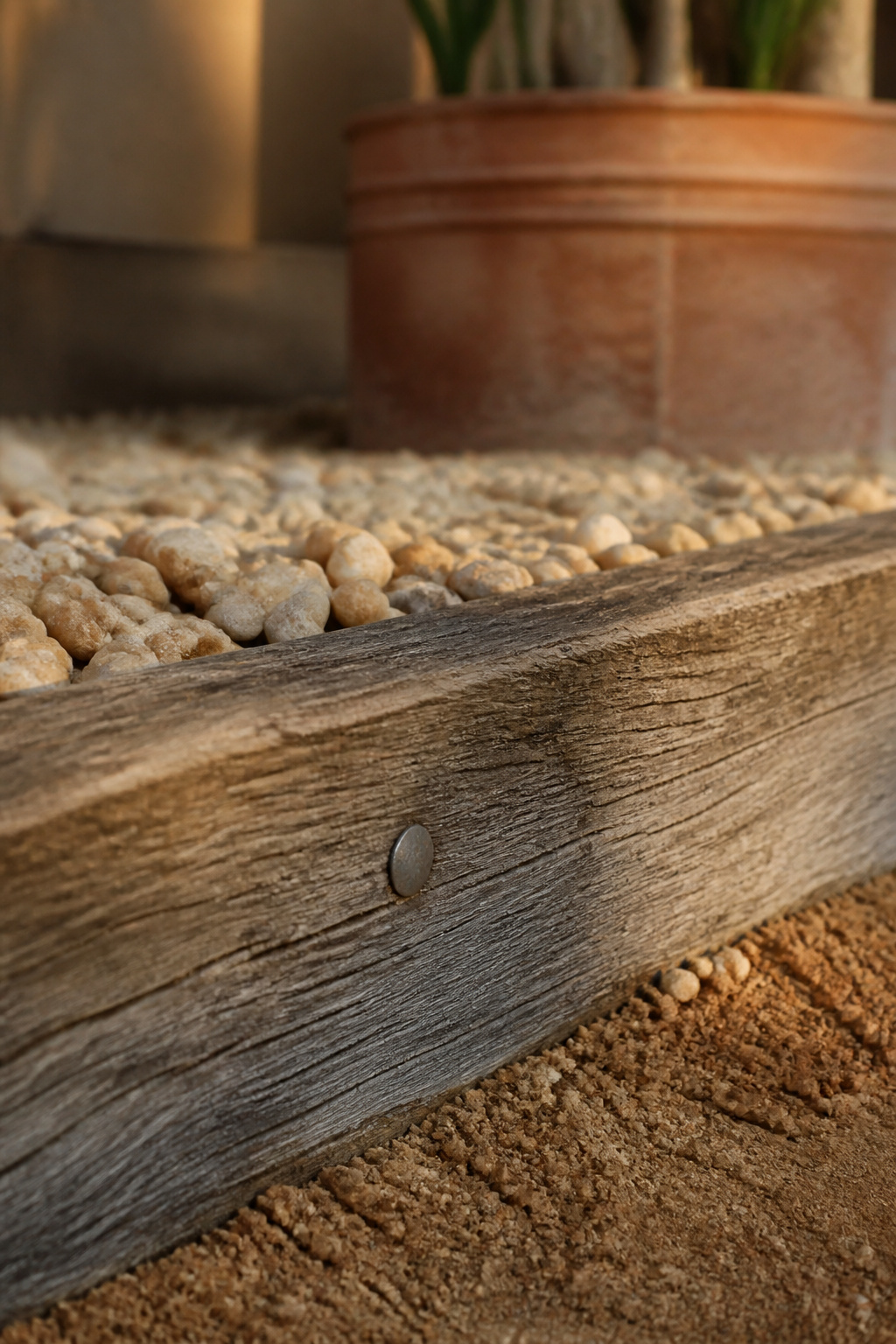 Weathered timber edging board bordering buff pea gravel with a terracotta pot nearby