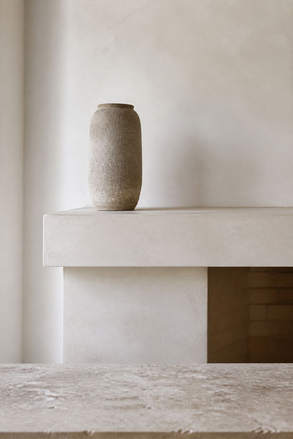 A pale plaster fireplace mantel with a single tall unglazed stoneware vessel, demonstrating minimalist mantel styling through negative space and material honesty