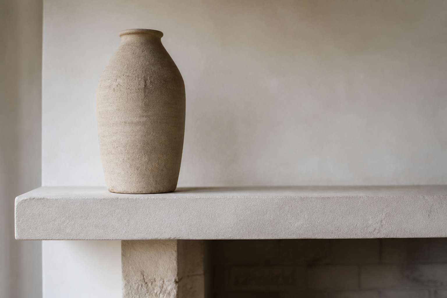 A pale plaster fireplace mantel with a single tall unglazed stoneware vessel, demonstrating minimalist mantel styling through negative space and material honesty