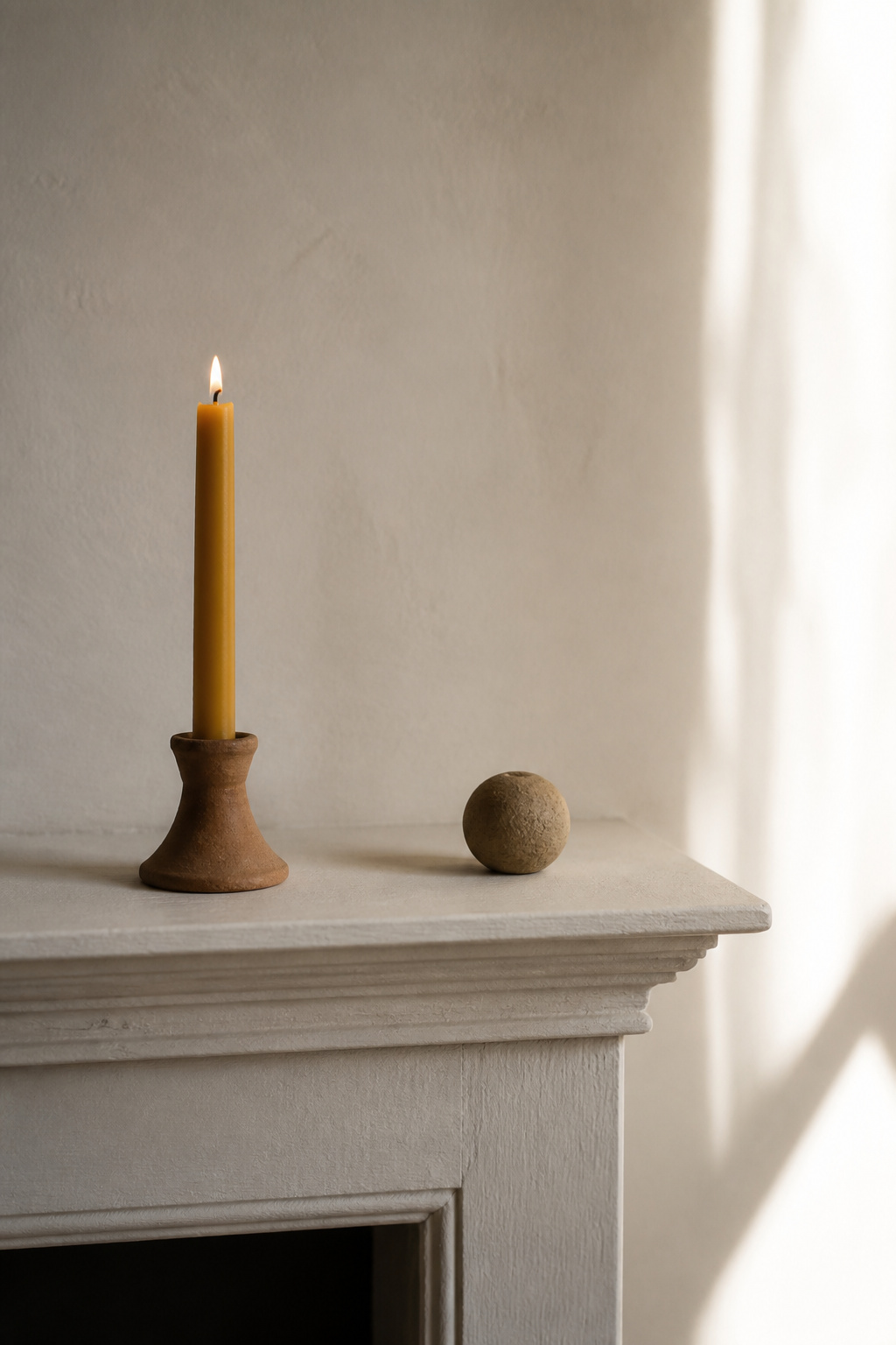 Two objects on a white minimal fireplace mantel — a beeswax candle in a clay holder and a stone sphere — illustrating material honesty and deliberate scale in minimalist styling