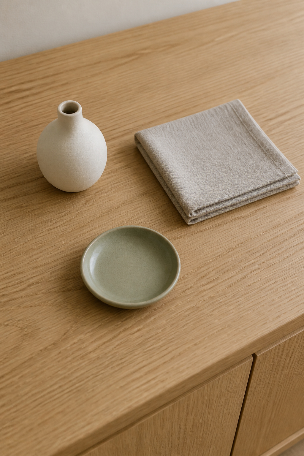 Pale oak sideboard with three palette tones — a warm white ceramic bud vase, a cool greige linen square, and the natural wood surface — plus one sage green accent dish, showing the tight neutral palette for modern sideboard styling