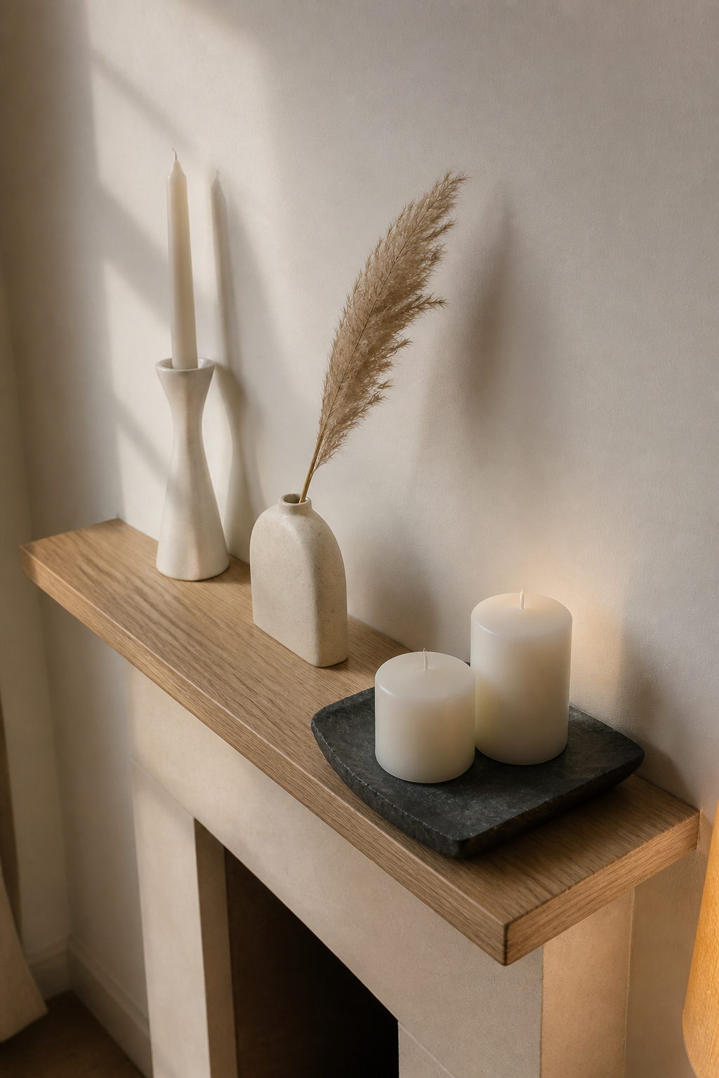 Shallow narrow mantel shelf with three objects in diagonal triangle: tall candlestick, bud vase with pampas, and low candle tray