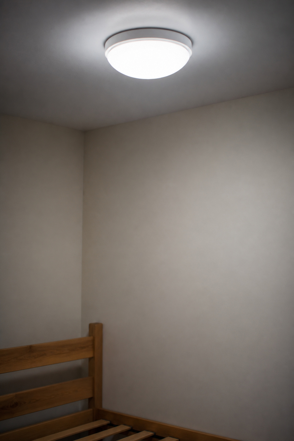 A plain bedroom corner lit only by a flat, cool flush-mount ceiling fixture with no other light source