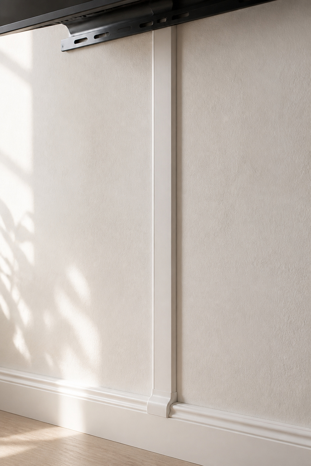 Colour-matched surface cable raceway running vertically on a warm white wall below a TV mount, nearly invisible against the wall