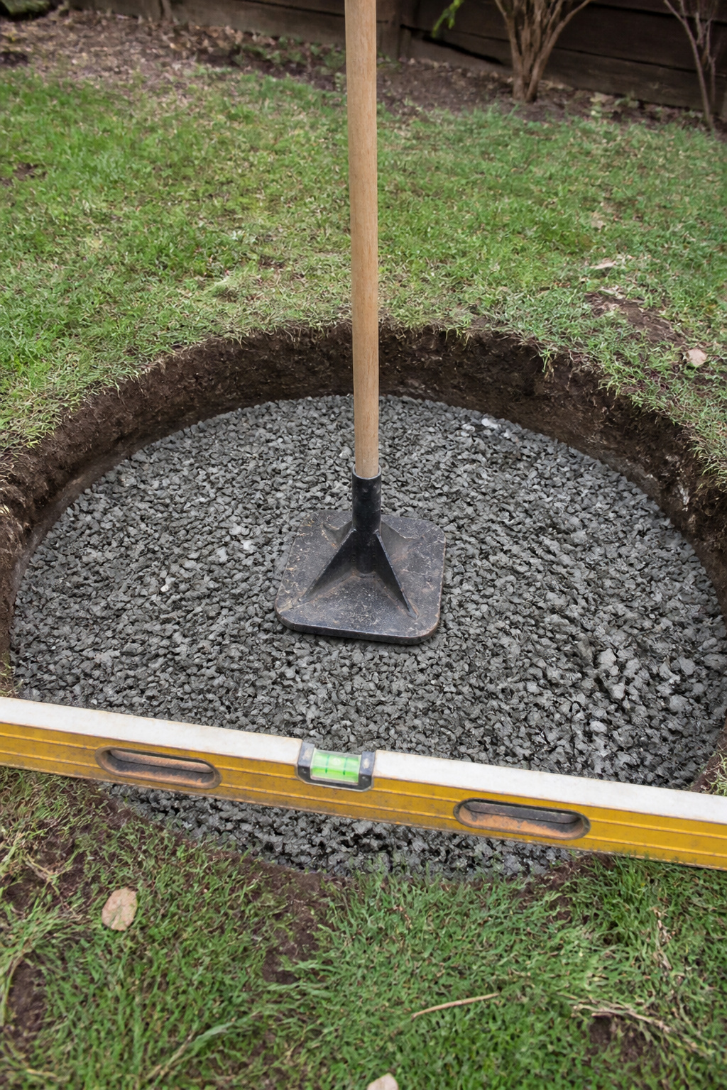 Excavated fire pit hole with compacted crushed gravel base layer and hand tamper mid-compaction, spirit level checking flatness