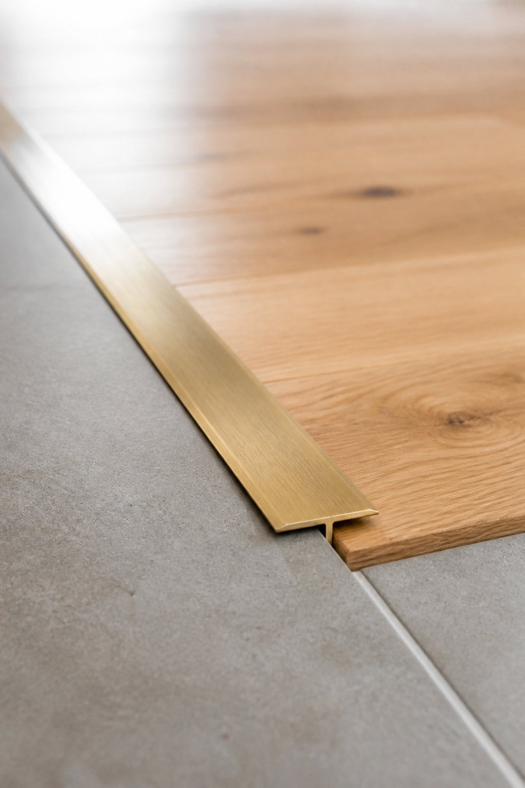 Slim brushed brass Schluter transition profile at the join between porcelain tile and white oak hardwood floor