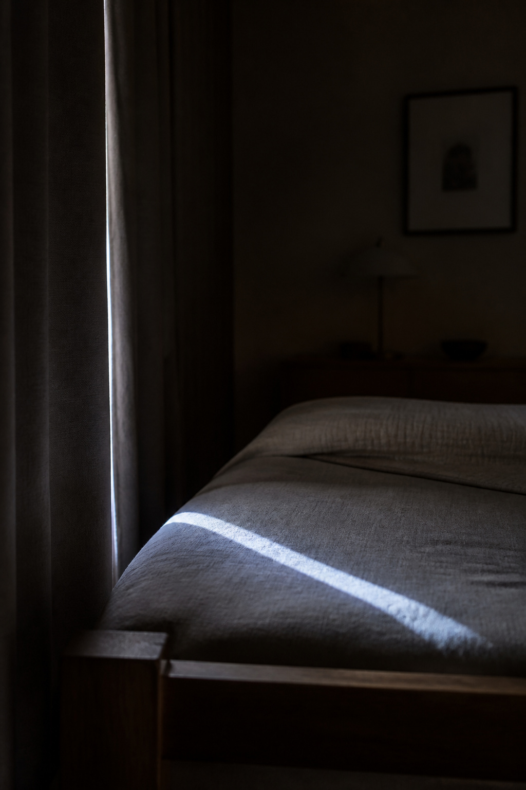 Thin sliver of streetlight crossing a dim bedroom at night through a gap beside the curtain