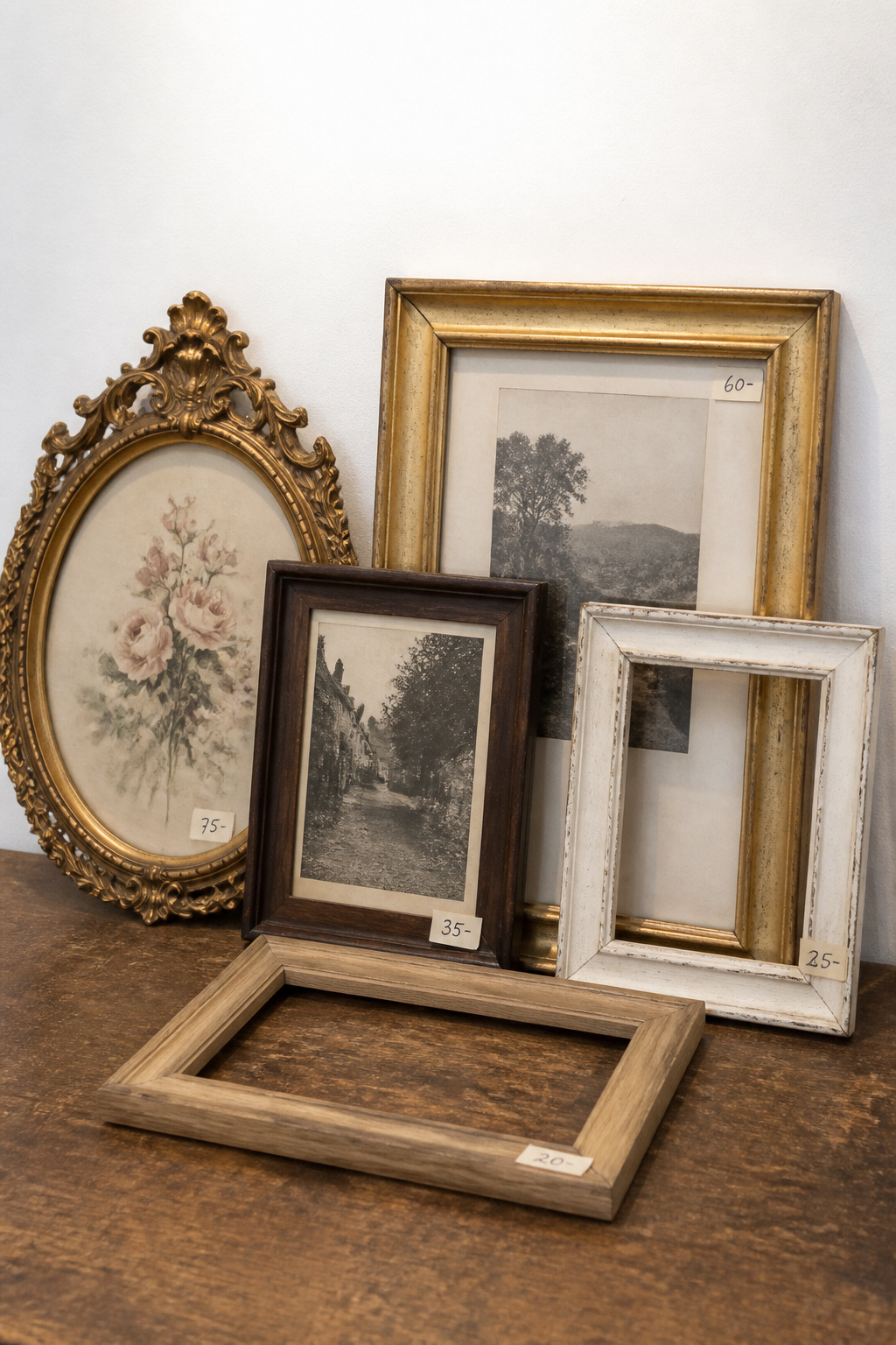 Five vintage frames leaning against a white wall on a table — gilt baroque oval, gilded rectangle, dark walnut, distressed cream and natural oak — some filled with art, two empty, as if displayed at an estate sale