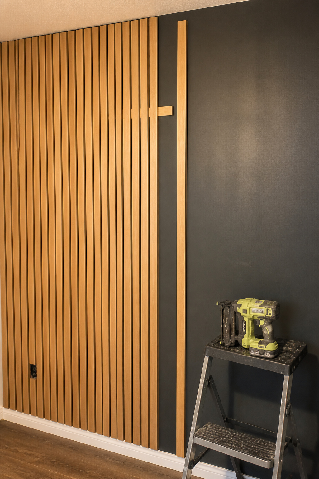Wood slat wall mid-installation showing spacer block technique and brad nailer, dark painted wall visible on right half
